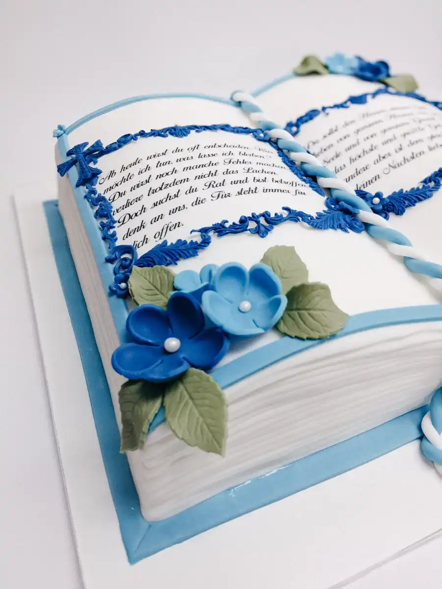 Festliche Buch-Torte zur Kommunion oder Konfirmation mit Fondant, blauen Blüten, Ornamenten und Textgestaltung.