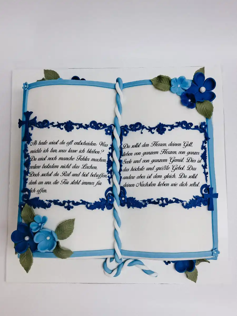 Weiße Buch-Torte zur Kommunion oder Konfirmation in Draufsicht, mit blauen Ornamentrahmen, geflochtenem Zuckerband und Blütendekoration.