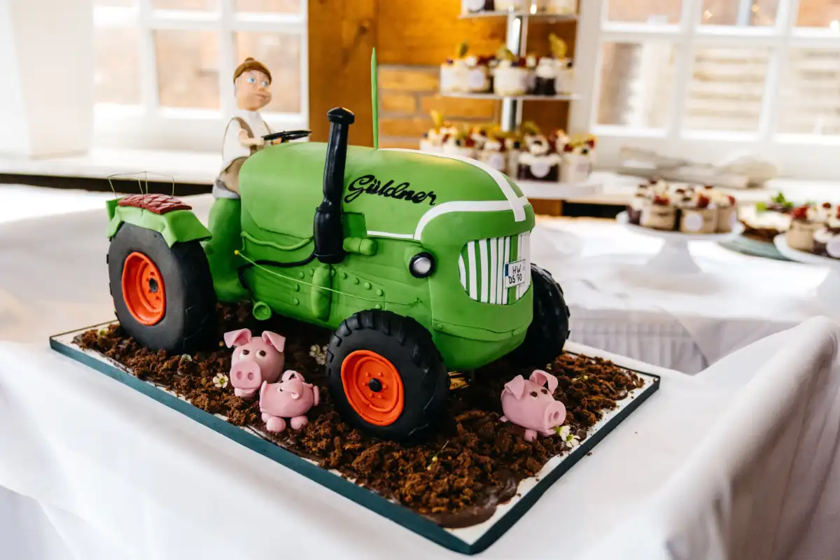 Motivtorte in Form eines grünen Güldner-Traktors mit modellierter Landwirt-Figur und kleinen rosa Schweinchen, handgefertigt von 7 Sachen Manufaktur.
