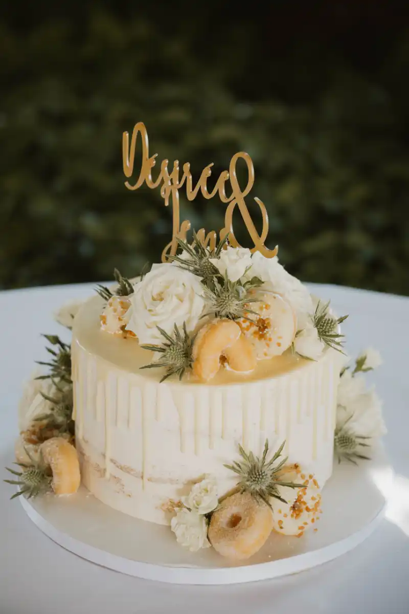 Hochzeitstorte mit Drip-Effekt und Blütendeko Einstöckige Hochzeitstorte mit Drip-Effekt, weißen Blüten, grünen Blättern, kleinen Donuts und goldenem Cake-Topper in Nahaufnahme.