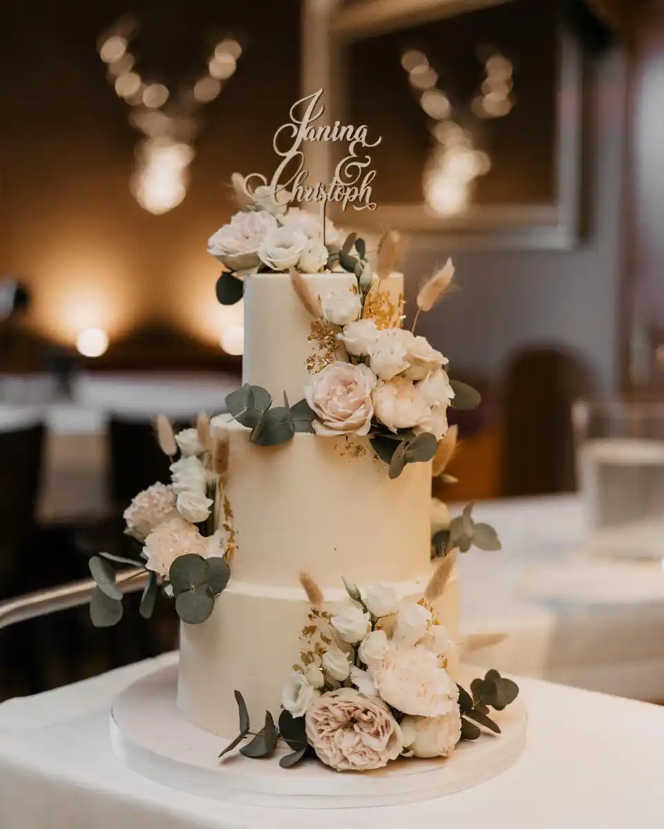 Dreistöckige cremefarbene Hochzeitstorte mit frischen Rosen, Eukalyptus und goldenen Akzenten, verziert mit personalisiertem Cake Topper für Janina & Christoph.