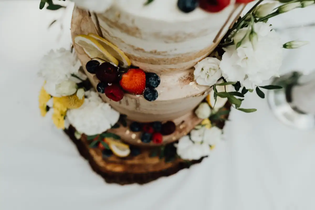 Detailaufnahme Dreistöckige Naked Cake Hochzeitstorte mit Beeren, Zitronenscheiben, weißen und gelben Blumen sowie personalisiertem Cake Topper auf Holzuntersetzer