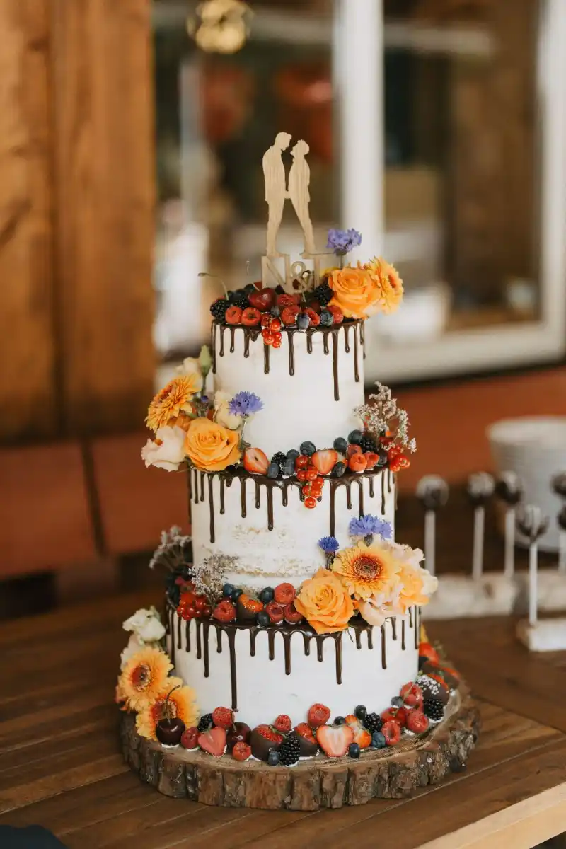 Dreistöckige Semi-Naked-Hochzeitstorte mit dunklem Schokodrip, dekoriert mit frischen Erdbeeren, Himbeeren, Brombeeren, Heidelbeeren, gelben und violetten Blüten sowie einem Cake Topper mit Brautpaar-Silhouette.