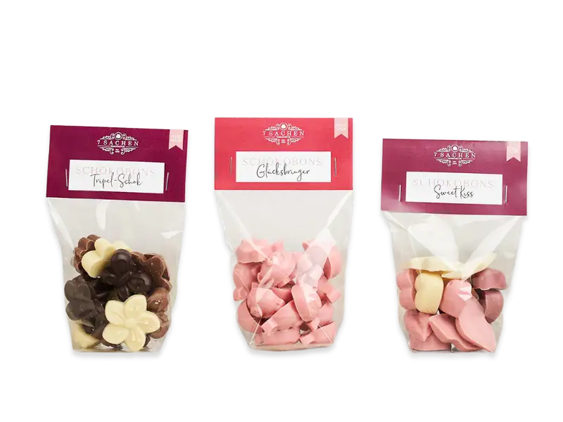 Drei Sorten handgemachte Schokobonbonss von 7 Sachen Manufaktur – Triple Schoki, Glücksbringer und Sweet Kiss – in transparenten Beuteln mit rotem Etikett.