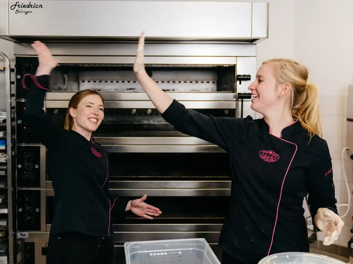 Zwei Konditorinnen geben sich in der Backstube ein High-Five und lächeln.